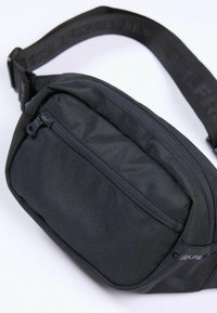 Geantă de talie din material textil negru, cu buzunar frontal cu fermoar și curea reglabilă, având text în relief pe un fundal alb.