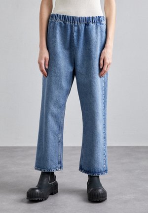 Personne portant un pantalon en jean bleu clair ample avec une taille élastique et des bottines noires épaisses, debout sur un sol gris.