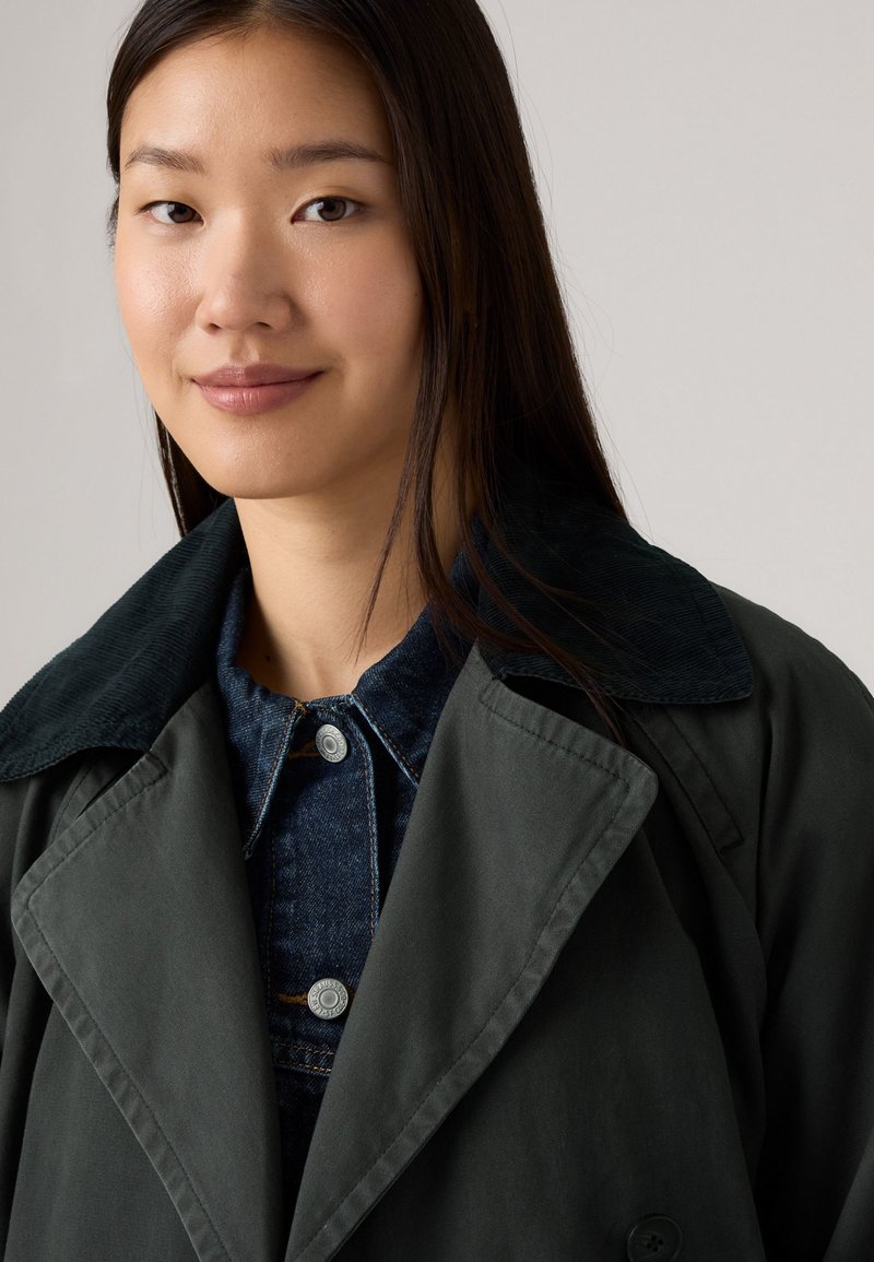 Manteau trench vert foncé avec un col en velours côtelé, superposé à une veste en denim dotée de boutons en argent. Texture lisse et design structuré.