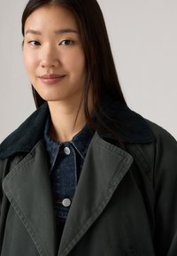 Mørkegrønn trenchcoat med en strukturert kordfløyelskrage, laget over en denimtop med trykknapper og klassisk krage.