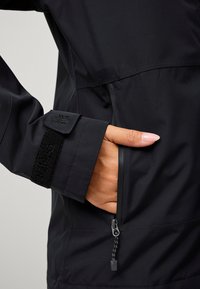 Giacca impermeabile nera con un design elegante, polsino in velcro testurizzato e tasca laterale con zip e cordoncino.