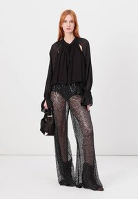 Zwarte, losvallende blouse met een trekkoordhalslijn, gecombineerd met doorschijnende, sprankelende zwarte wijde broek. Het model houdt een kleine handtas vast.