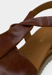 Καφέ δερμάτινο σανδάλι με σχέδιο στριφτού λουριού, λεία υφή και ταμπά εσωτερική σόλα με ανάγλυφη επιγραφή.