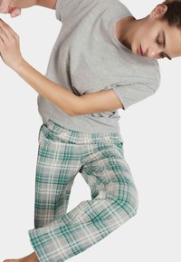 Joven en camiseta gris y pantalones de cuadros verdes estirando el brazo con los ojos cerrados sobre un fondo claro.