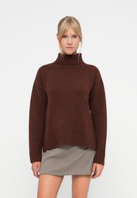 Bruine oversized trui met hoge hals, met geribbelde manchetten en zoom. Gelaagd textiel met een subtiel breipatroon. Gecombineerd met een geruit rokje.