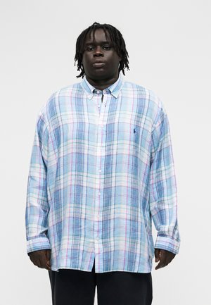 Vyras su tamsia oda ir dredi plaukais dėvi permatomą mėlyną, baltą ir rožinę languotą marškinių stilių su juodomis kelnėmis, stovintis prieš baltą foną.