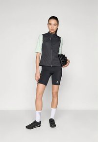 Μαύρο γιλέκο με φερμουάρ, ανοιχτό πράσινο κοντομάνικο μπλουζάκι, μαύρα σορτς ποδηλασίας, λευκές κάλτσες, μαύρα παπούτσια ποδηλασίας· κράνος κρατημένο σε ένα χέρι.