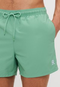 Șorturi de baie verde mentă cu talie elastică, șnur, buzunare laterale și un mic logo alb pe tiv. Textură de material neted.