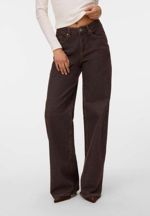 Femme portant un jean marron foncé taille haute à jambes larges et un haut blanc à manches longues, tenant l'ourlet du haut près de la taille.