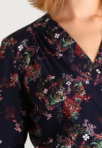 Marinebluse mit floralen Mustern, die rote, rosa und grüne Blumen zeigt. V-Ausschnitt und gerafftes Gewebe an den Schultern. Weicher Stoff.