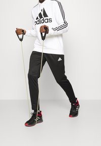 Bílá mikina Adidas s černým logem, černé kalhoty s bílým logem Adidas, černé a červené sportovní boty, držení odporových gum.