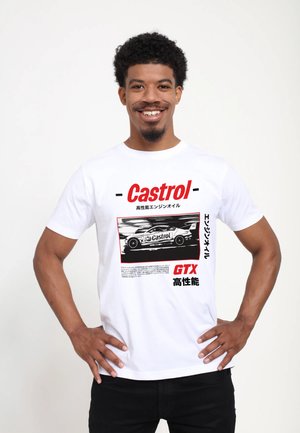 Homme souriant aux cheveux bouclés portant un t-shirt blanc Castrol GTX avec une voiture de course noir et blanc et un texte japonais rouge, les mains sur les hanches.