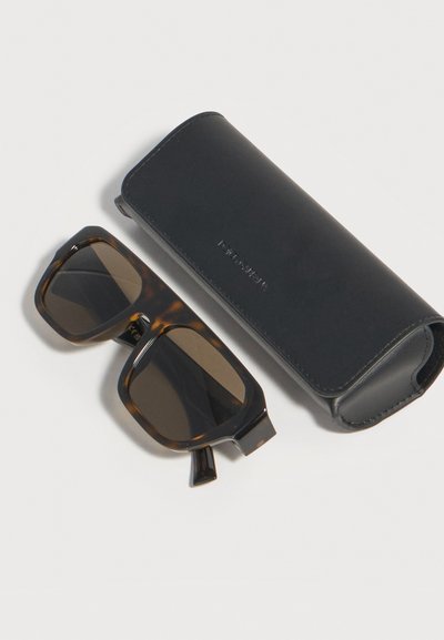 Τετράγωνα γυαλιά ηλίου σε σχέδιο χελώνας με σκούρους φακούς δίπλα από μαύρη θήκη γυαλιών ηλίου από δέρμα πάνω σε ανοιχτόχρωμη επιφάνεια.