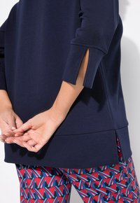 Maglione blu navy con maniche leggermente svasate e spacchi laterali, abbinato a leggings con fantasia che presentano design geometrici in rosso, blu e bianco.