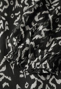 Schwarz-weiß gemusteter Stoff mit abstrakten Designs. Das Material wirkt leicht mit einer strukturierten, gerüschten Oberfläche und einem gerafften Ärmelabschluss.