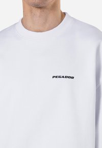 Sudadera blanca con un cuello redondo acanalado y un logo de texto negro "PEGADOR" en el lado izquierdo del pecho. Tejido suave con una textura lisa.