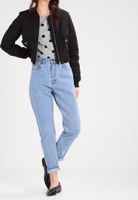 Kvinna i ljusblå högmidjade jeans, svart bomberjacka, grå skjorta med prickar och svarta slip-on skor, stående med ett ben korsat.