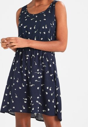 Kobieta w granatowej sukience bez rękawów z drobnym białym kwiatowym wzorem, stoi ze złożonymi rękami przed sobą.