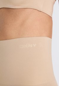 Top scurt DKNY bej, cu o textură netedă și o bandă lată cu numele mărcii subtil embosat. Vedere de aproape a țesăturii și a ajustării.