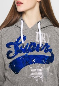 Gray hooded sweatshirt with blue sequin text "Super," white drawstrings, and silver star graphic. Textured, shiny fabric with a sparkling effect.