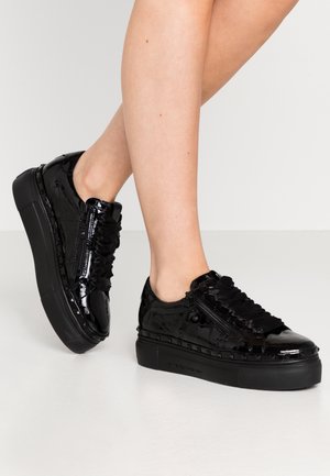 Nogi w czarnych lakierowanych trampkach na platformie z marszczonymi sznurowadłami i bocznymi zamkami, stojące na gładkiej jasnej podłodze.