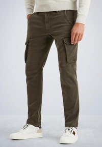 Pantalons cargo vert olive avec plusieurs poches latérales, détails zippés et coupe slim ; assortis à des baskets blanches avec des accents noirs et beiges.