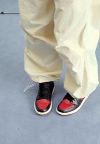 Persona che indossa pantaloni beige larghi e sneakers nere, rosse e bianche, in piedi su un pavimento grigio chiaro.