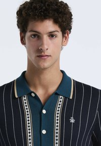 Marine gestreepte polo met een teal kleurige kraag, drie knopen en accenten in diamantpatroon. Heeft een klein logo aan de linkerkant.