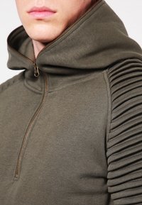 Olivgrön zip-up hoodie med hög krage och ribbad ärmadetalj. Tillverkad av mjukt tyg med slät textur. Gulddragkedja synlig.