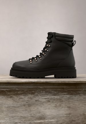 Bottine en cuir noir avec semelle épaisse à crampons, œillets métalliques, col matelassé et lacets à l’avant sur surface en bois sur fond neutre.