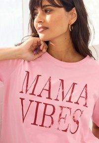 Roze katoenen T-shirt met de opvallende tekst "MAMA VIBES" in versleten rode letters. Korte mouwen en een ontspannen ronde hals.