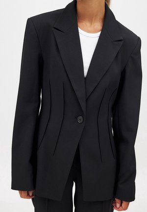 Kvinde iført en tætsiddende sort blazer med en enkelt knap over en hvid ribbet top, stående mod en ensfarvet lys baggrund.