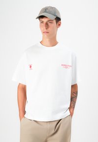 White cotton t-shirt with round neckline, short sleeves, and pink printed branding on the left chest. Paired with beige trousers and a faded cap.