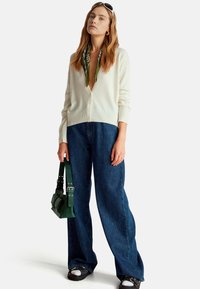 Bílá pletená kardigán přes zelený šátek, v kombinaci s širokými modrými džínami a malou zelenou kabelkou. Černé tenisky dotvářejí vzhled.