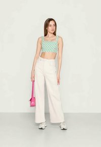 Jovem mulher vestindo calças largas brancas, um top cropped verde com bolinhas, ténis brancos, segurando uma pequena bolsa rosa brilhante, em pé sobre um chão cinza.