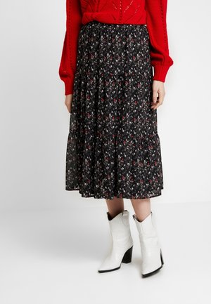 Une femme porte une jupe midi noire à fleurs, un pull rouge à manches longues, et des bottes blanches à talons mi-mollet, debout devant un fond blanc.