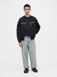 Jeune homme portant une veste noire zippée avec un texte blanc, un jean large bleu clair et des baskets noires, debout devant un fond blanc uni.