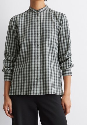 Person iført en langærmet, grå og sort ginghamternet skjorte med opkrullede ærmekanter og sorte bukser, stående mod en hvid baggrund.