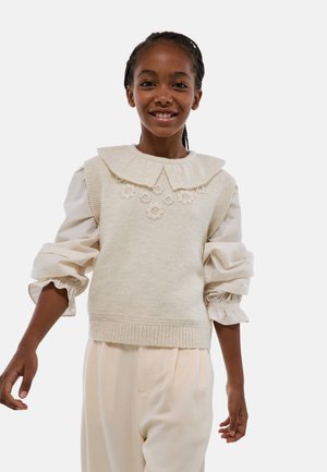 Pull en maille crème avec un col Peter Pan et des broderies florales, associé à une blouse crème clair avec des manches volumineuses et volantées.