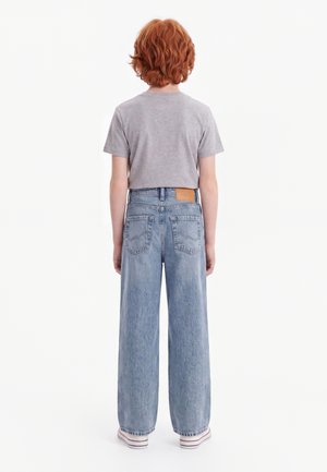 Barn med krøllet rødt hår står med ryggen mod iført en grå t-shirt, blå vidde bukser og hvide sneakers mod en ensfarvet hvid baggrund.