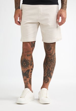 Pantalones cortos beige de algodón con corte recto, que cuentan con bolsillos laterales y un puño con dobladillo, combinados con zapatillas blancas.