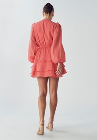 Frau mit einem Dutt, die ein korallenfarbenes langärmliges gestuftes Minikleid und nude-farbene High-Heels trägt, steht vor einem hellen, einfarbigen Hintergrund.