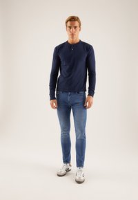 Cămașă bleumarin cu mâneci lungi și decolteu henley, asortată cu blugi slim fit bleu deschis și adidași gri cu dungi negre.