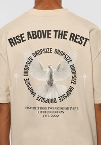 Beige bomulds t-shirt med en sort grafik af en due med udbredte vinger, omgivet af teksten "REJS DIG OVER DE ANDRE" og "DROPSIZE."