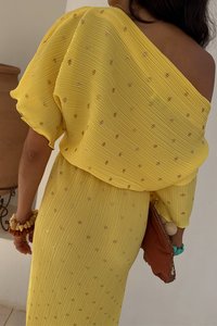 Geel, geribbeld jurk met een off-shoulder ontwerp, voorzien van gouden sequins. Vergezeld van een bruine clutch en kleurrijke accessoires.