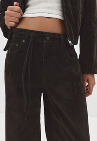 Mustat corduroy-housut rennolla istuvuudella, joissa on nyöri-vyötärö, erottuvat siniset tikkaukset ja etutaskut. Sujuva pinta.