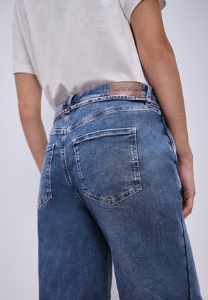 Person trägt blaue Jeans und ein weißes Hemd, von hinten gesehen, mit Fokus auf die Taschen- und Bunddetails der Jeans.