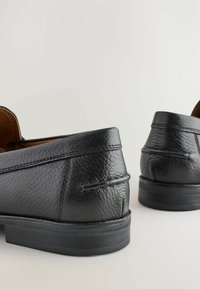 Mocasines de piel negra con un acabado texturizado, tacón bajo y un interior marrón en contraste. Presentan detalles de costura a lo largo de los bordes superiores.