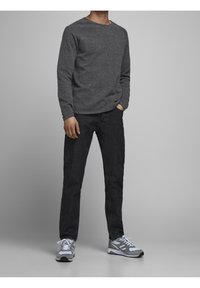 Mężczyzna stoi z rękami w kieszeniach, ubrany w ciemnoszary długi rękaw, czarne dżinsy i szaro-białe sneakersy na neutralnym tle.