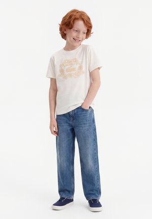 Enfant roux souriant, debout avec une main dans la poche, portant un t-shirt blanc Lacoste, un jean bleu et des baskets bleu marine.